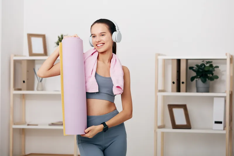 Female fitness enthusiast in athletic wear with headphones holding a pink yoga mat in a bright home gym, ready for workouts, stretching and strength training.