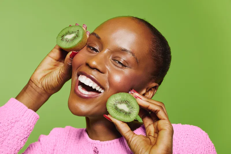 Happy African woman with dark skin holds two halves of fresh kiwi near face, smiling joyfully on green background. Concept of healthy lifestyle, natural beauty, and fruit wellness.