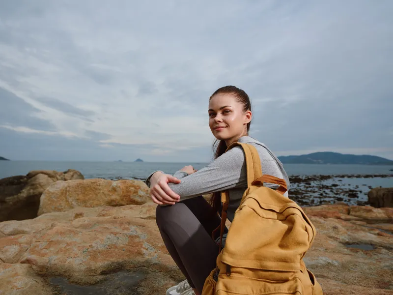 Young traveler with backpack sitting on a rocky shore, facing the sea Embracing adventure and exploration under a moody sky, ideal for travel and lifestyle themes