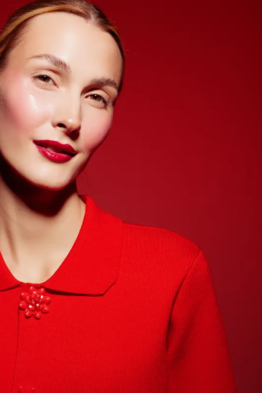 Portrait of a confident woman with red lipstick and red blouse against a red background. Studio lighting highlights her glowing skin and elegant expression. Concept of beauty and fashion.