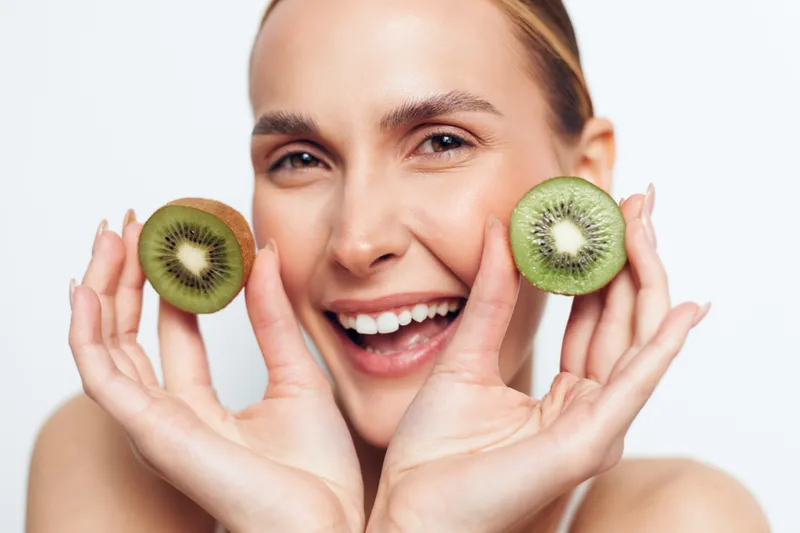 A smiling young woman holds two kiwi halves near her cheeks, creating a playful fruit punch pose that communicates fresh skincare and healthy eating in a bright studio setting.