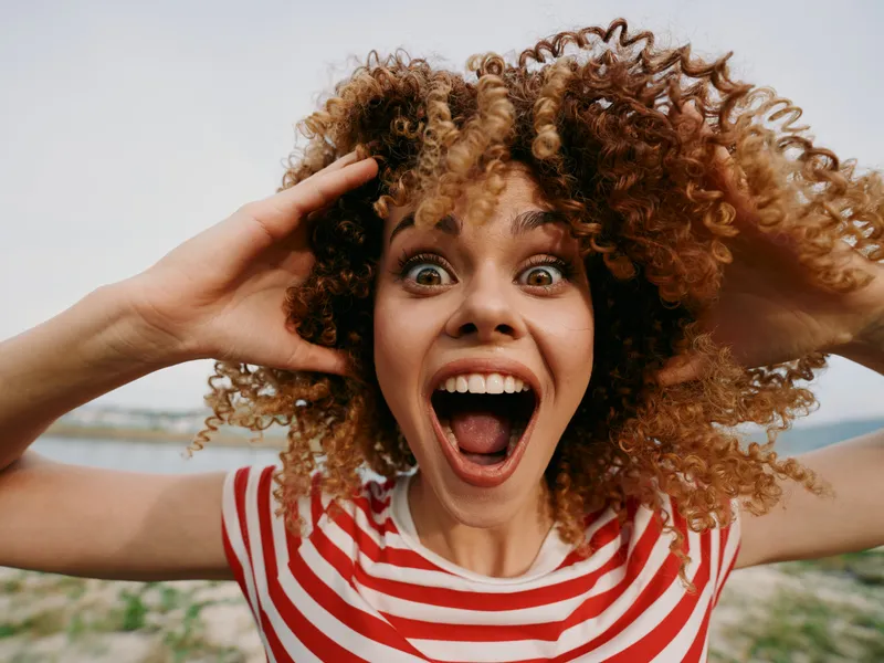 Vibrant portrait of a young woman with curly hair at the beach, eyes wide and mouth open in surprise, wearing a striped shirt and exuding energetic joy.
