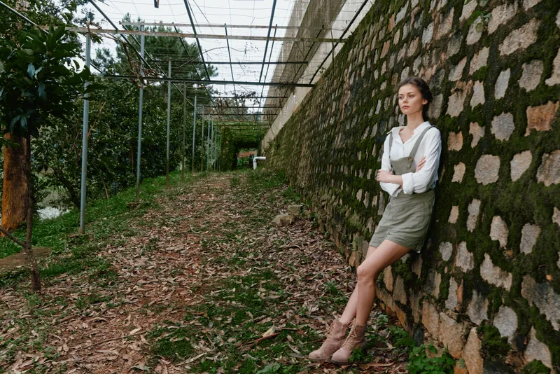 Young woman in casual green overalls poses confidently against a rustic stone wall in a lush garden, exuding a relaxed and serene vibe