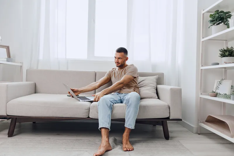 Focused man working on a laptop in a cozy living room, adorned with greenery and soft natural light, captures a modern, tranquil lifestyle.