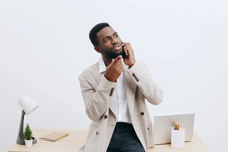 Thoughtful young black man in beige blazer and white shirt sitting at modern minimalist desk with laptop, notebook, and plant, posing in bright neutral office space. Business casual look, contemplativ