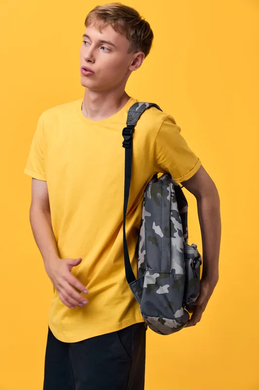 Backpack wearing youth in casual attire with a camo backpack standing against a bright yellow backdrop, ready for school, daily commute, or weekend adventure and spontaneous exploration, showcasing