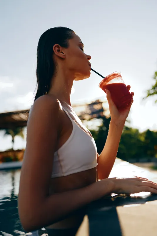 Woman lounges by a sunlit pool, sipping juice through a straw, enjoying a relaxed summer moment by the water, showcasing outdoor lifestyle and tranquil poolside ambiance
