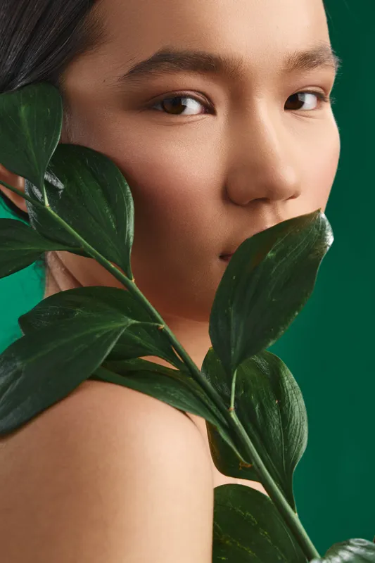asian woman with a calm expression is partially obscured by lush green leaves in a vibrant studio setting, conveying natural elegance and serene beauty.