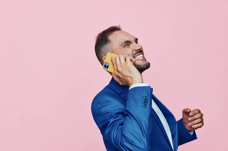 A professional man in a blue suit speaks on a yellow phone against an isolated pink background, conveying energy, focus, and bold business intent.