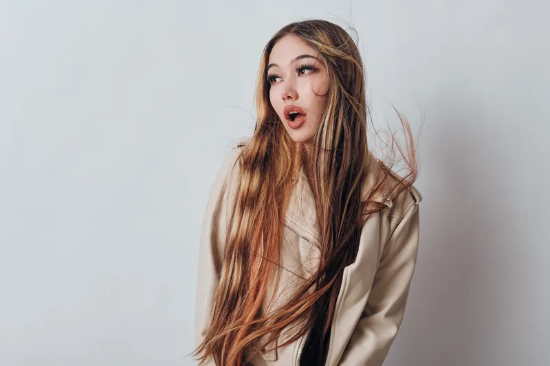 Young woman with long hair wearing casual light jacket on neutral background looking surprised with open mouth and expressive face, hair slightly moving in the air.