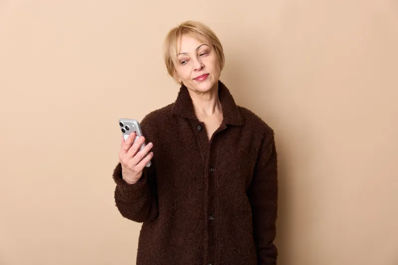 Woman with smartphone, portrait indoors, smiling and looking at screen, using mobile communication technology, warm beige background, casual coat, calm expression today
