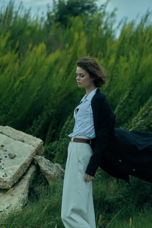 Woman in a windblown coat stands in a grassy field, white shirt and dark blazer contrasting with open landscape, creating a cinematic mood and subtle motion.