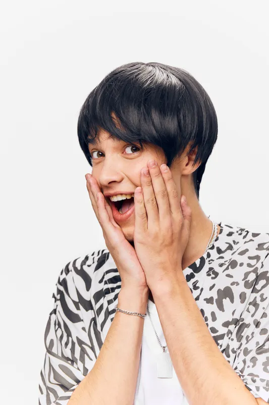 surprised young man with short black hair wearing patterned shirt showing excited expression with hands on cheeks against white background, casual style