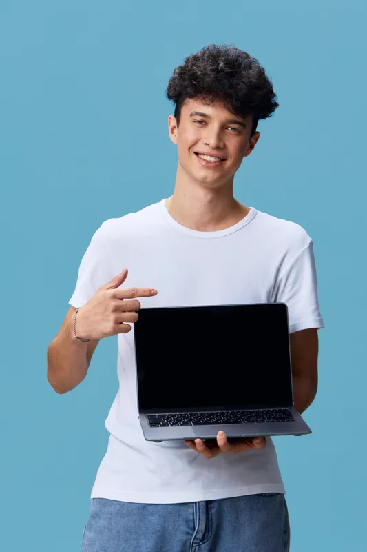 smiling man holding laptop and pointing at screen, casual young adult wearing white t-shirt on blue background showing digital device