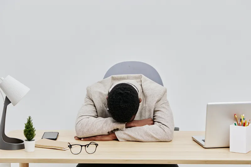 Businessman with dark hair and beard in beige sweater sitting at desk with head down in hands, surrounded by laptop, glasses, smartphone, and stationery, expressing stress or frustration, neutral back