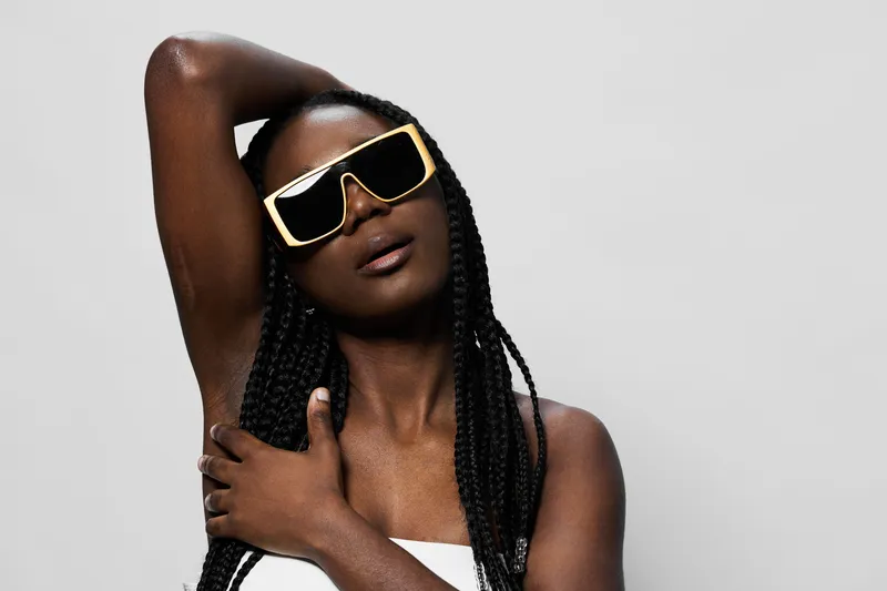 Elegant African woman wearing trendy sunglasses poses confidently, showcasing her braided hair and stylish outfit against a neutral background