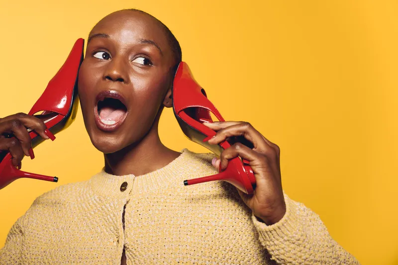 Surprised black woman with dark skin holds red high heels near her face against yellow background. Concept of fashion, style, beauty, and expressive emotions in studio portrait.