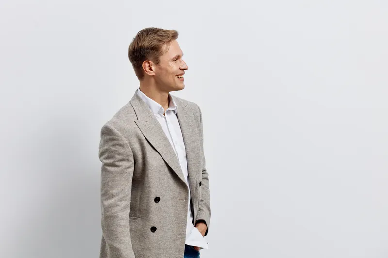 Confident man in stylish gray patterned blazer and white shirt standing against plain light gray background. Fashionable, professional appearance, business casual style, smiling and looking to the sid