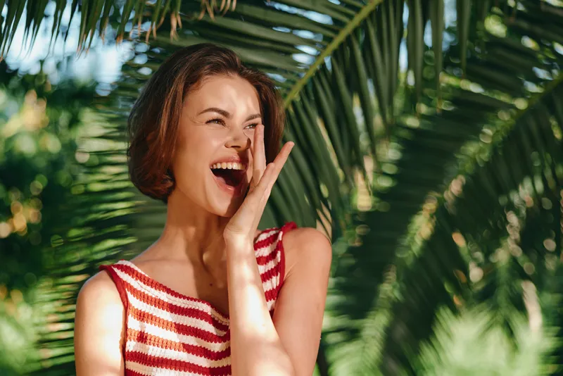A cheerful woman is laughing outdoors in a sunny garden, smiling broadly and posing candidly, creating a vibrant, relaxed summer mood with warm light and natural charm.