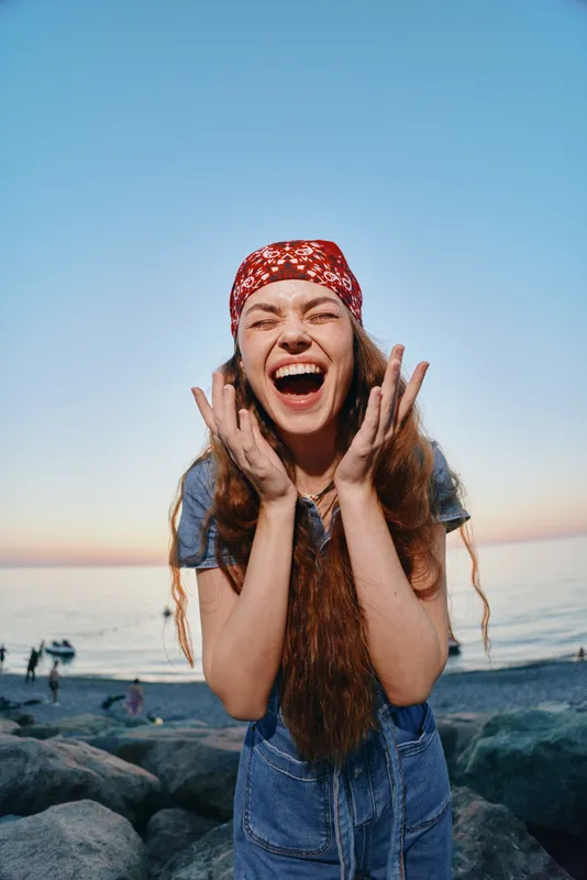 Lifestyle woman wearing modern boho-western denim street style with a red bandana, laughing joyfully by the rocky beach at sunset in warm film colors.