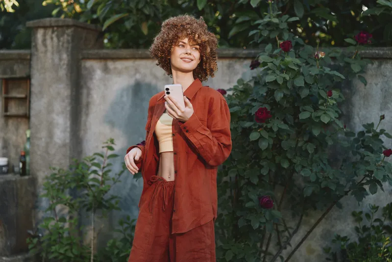 A woman in burnt orange outfit stands outdoors with a smartphone, smiling. Lush greenery and a garden backdrop create a bright, relaxed mood, highlighting casual fashion and confident lifestyle