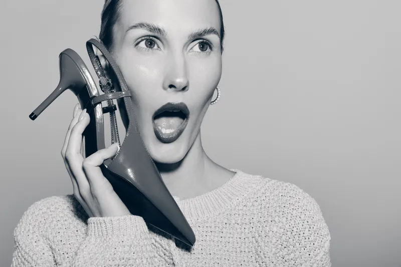 Surprised white woman with fair skin holds high heel shoe near face, wearing sweater in black and white studio photo. Concept of fashion and expression.