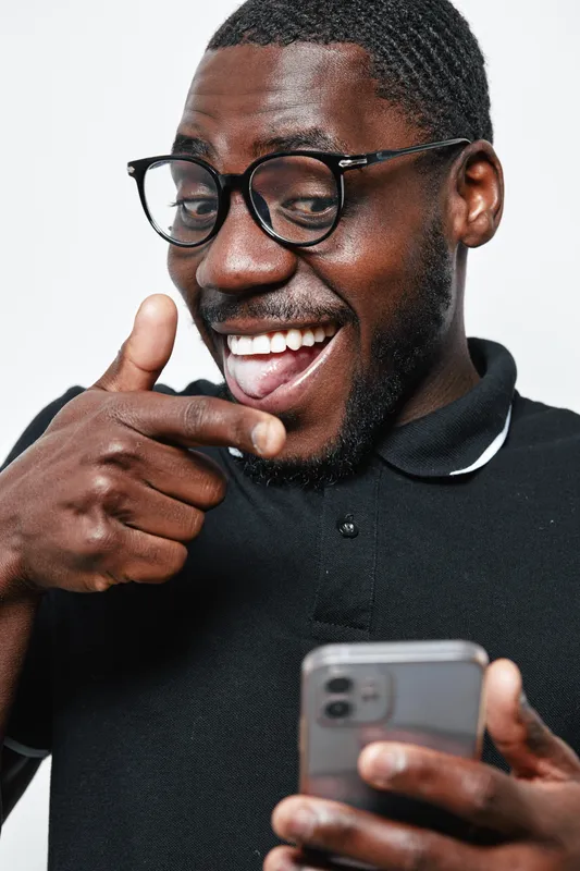 African American man with dark skin and glasses smiling and pointing finger while holding smartphone in casual black shirt on white background. Positive emotion and technology.