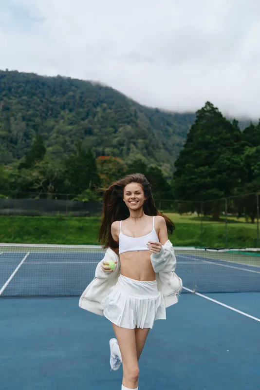 Young athletic woman running on a tennis court, dressed in a white sports outfit, embodying joy and energy against a lush green backdrop