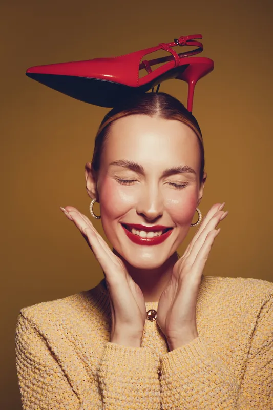 Smiling woman with red high heel shoe on head poses with closed eyes and hands near face. Fashion concept with playful mood and bright studio lighting on yellow background.