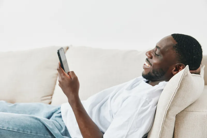 Smiling African man relaxing on sofa while using smartphone, enjoying leisure time at home, casual outfit, bright and airy background for relaxation concepts