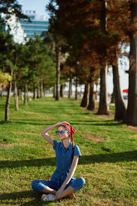 Lifestyle woman in modern boho-western denim street style sitting on grass with sunglasses and red headscarf, bright film color capturing relaxed urban vibe outdoors.