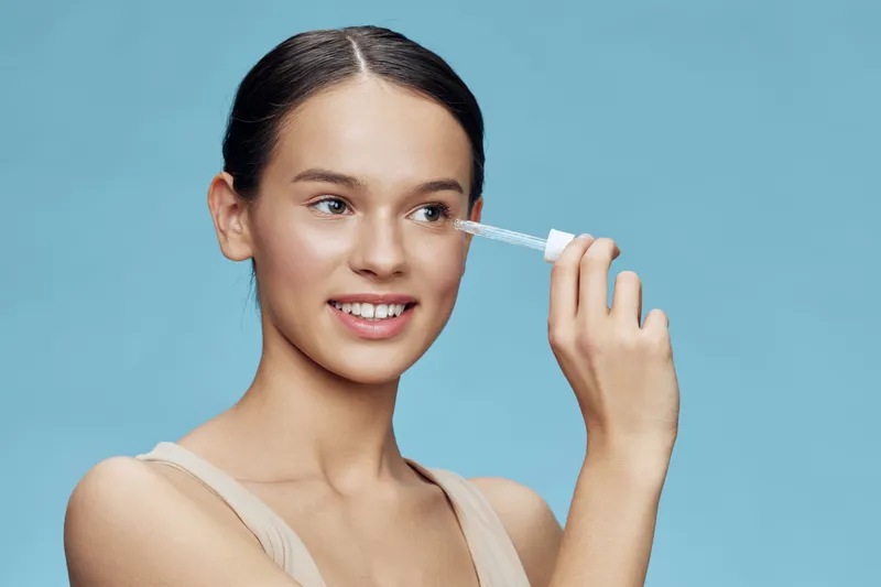 Young female model applying skincare serum with a dropper against a soft blue background, embodying fresh beauty and self care in a vibrant, serene atmosphere