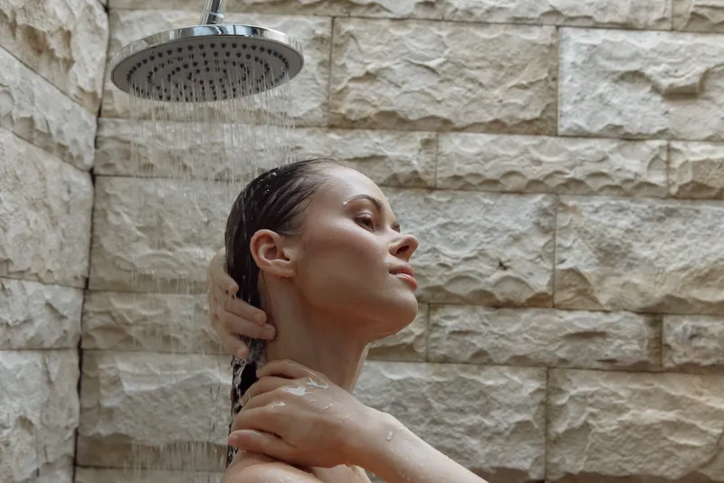 woman showering under rain showerhead with stone wall background, wet hair, relaxed expression, natural skincare routine, minimalistic bathroom space