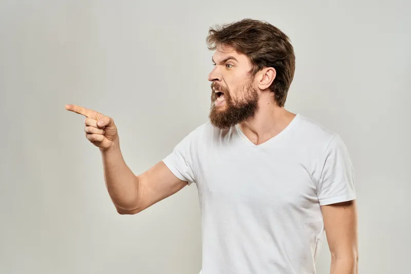 Бородатый мужчина в белой футболке жестикулирует руками на светлом фоне, его поза и выражение лица передают агрессию