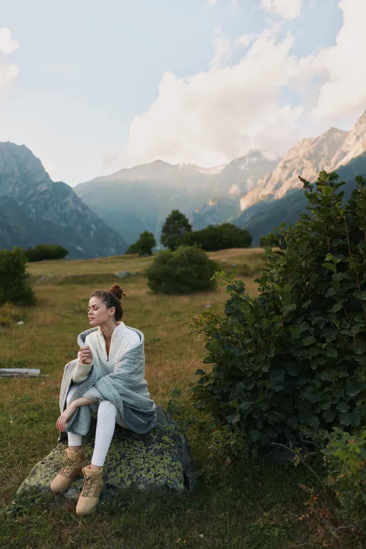 Young woman wrapped in blanket sitting on rock in mountain meadow holding cup. Relaxing outdoors with scenic landscape and soft natural light. Concept of lifestyle and calm moment.