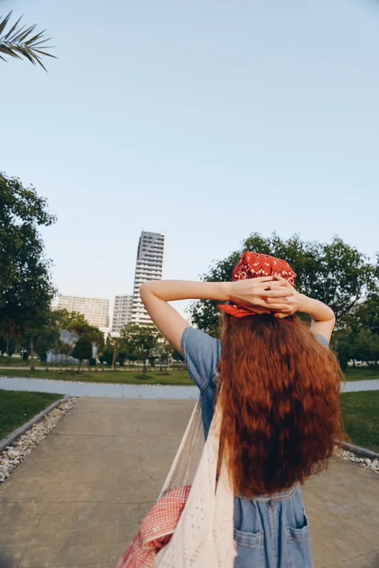 Lifestyle woman in modern boho-western denim street style with long red hair and bandana, enjoying outdoor urban park in film color tones during daytime.
