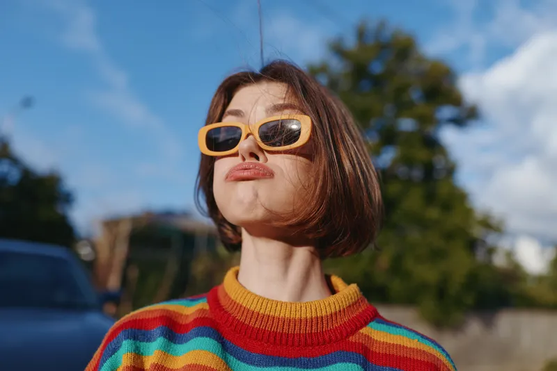 Woman young woman in her 20s wearing a striped sweater and yellow sunglasses, playful expression with a pout in an outdoor portrait on a sunny suburban street, lifestyle fashion travel mood.