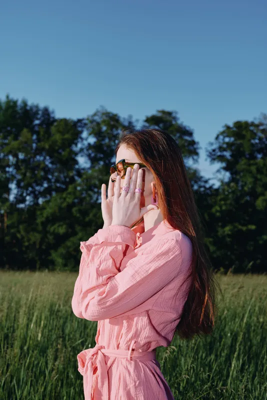 Stylish young woman in a pink dress covering her face with hands in a sunny field, capturing beauty and tranquility of summer in nature