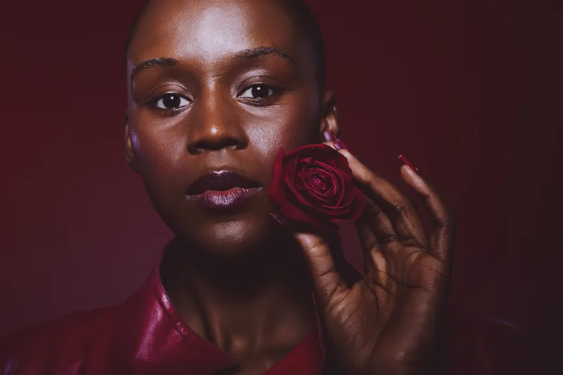 Close-up of a confident dark skinned African woman holding a red rose near her face. The studio portrait features glossy skin and deep red tones, symbolizing beauty and elegance.