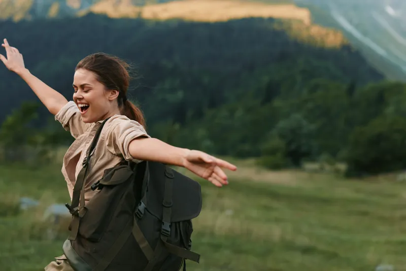 Young woman with a backpack explores a grassy hillside, arms wide, smiling at the open sky.