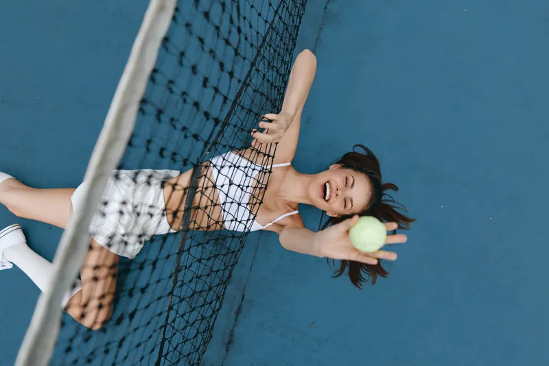 Joyful young woman enjoying a tennis game, lying on the court with a ball, vibrant blue background, perfect for sports and active lifestyle concepts
