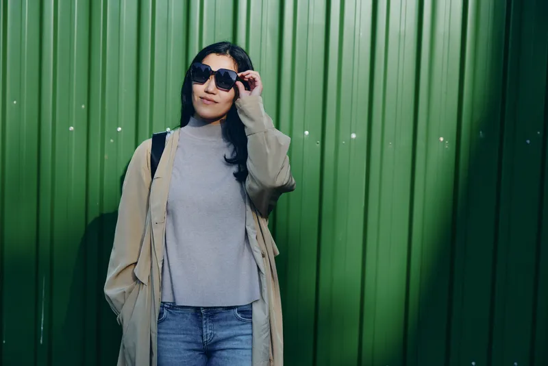 Woman wearing a beige trench coat, gray sweater and jeans stands against a green wall, adjusting sunglasses. Street style with a relaxed pose, urban backdrop, fashion vibe, minimal accessories and