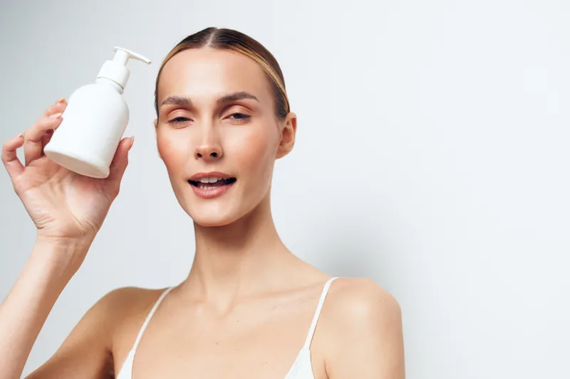 A smiling woman demonstrates a skincare routine, holding a white lotion bottle against a bright neutral background, emphasizing hydration, smooth skin, and daily self care.