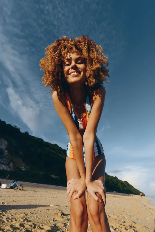 Smiling young woman with curly hair sitting on a sunny beach holding her knees, relaxed vacation mood, warm sunlight, blue sky and ocean horizon, outdoor lifestyle concept for summer joy and freedom