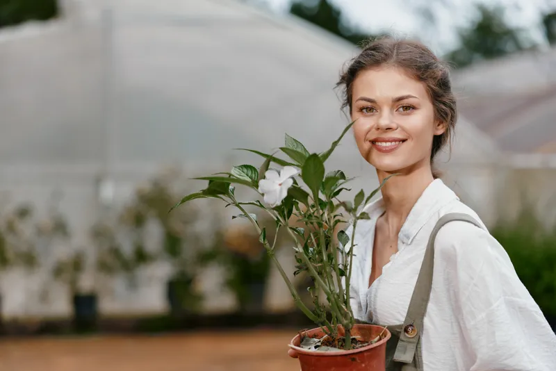 Young woman in a greenhouse holding a potted flower, showcasing a smile, dressed in a light blouse with natural colors, embodying joy and nature
