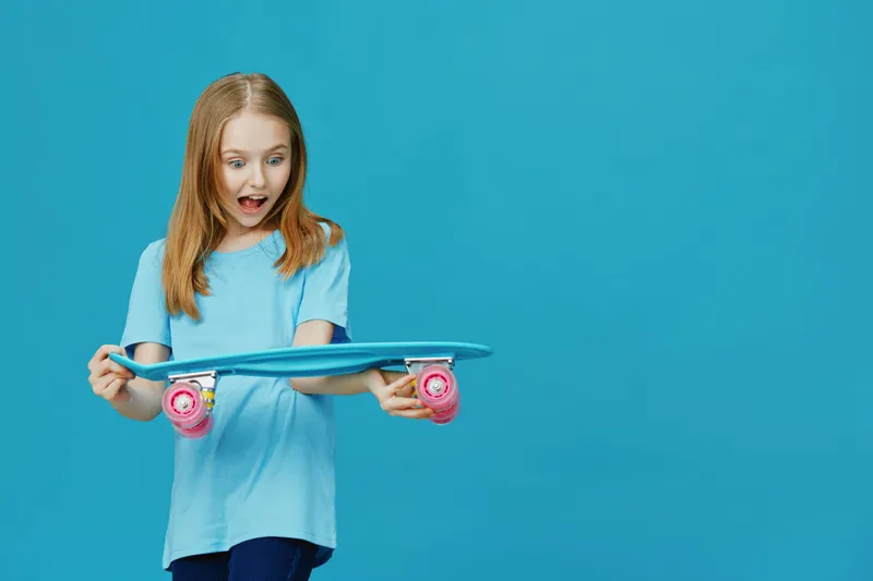 Excited girl with skateboard on a bright blue background, showcasing joy and surprise in a vibrant, youthful setting