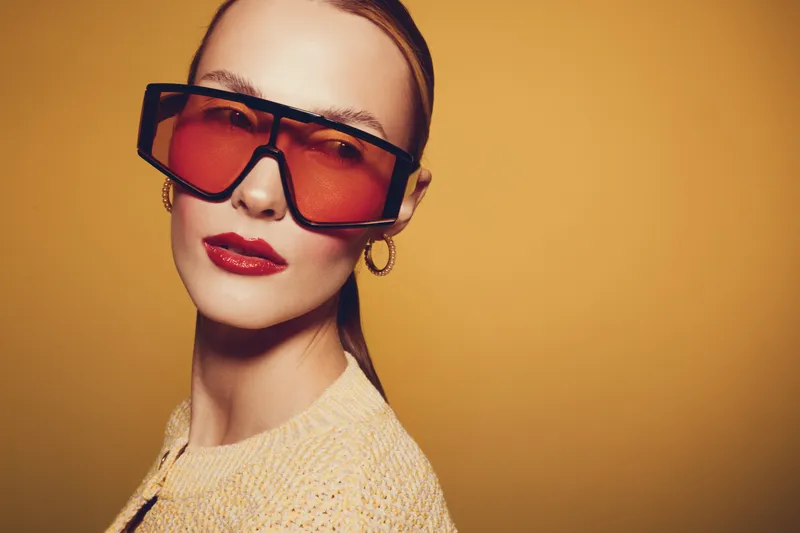 Stylish woman with red tinted oversized sunglasses and red lipstick poses against yellow background. Fashion portrait with modern accessories and confident expression in studio lighting.