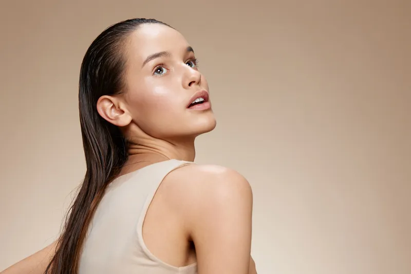 Confident young woman with long hair looking back, showcasing healthy skin and subtle makeup against a neutral background