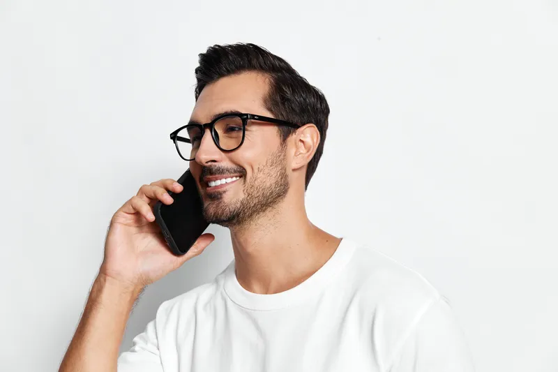 Smiling man wearing black glasses speaks on mobile phone with cheerful expression, isolated on clean white background. Modern lifestyle concept.