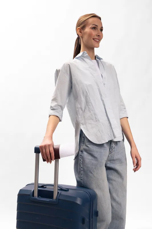 Young woman in casual clothes stands with suitcase and travel mug. Concept of travel, lifestyle, and modern trip preparation in minimal studio setting with white background.